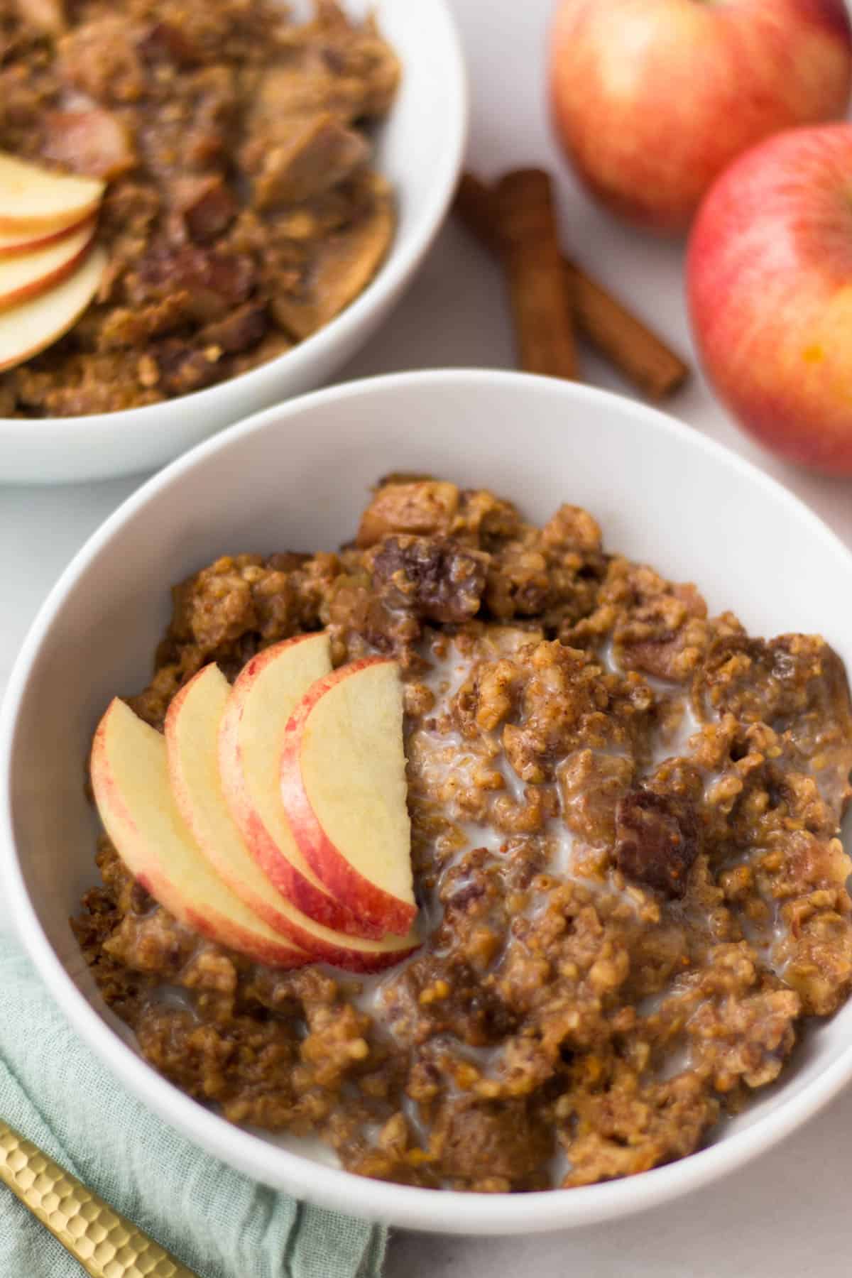 Close up of a bowl of these healthy Fig Apple Cinnamon Slow Cooker Steel Cut Oats topped with apples. Behind it is another bowl with oats, two whole apples, and cinnamon sticks.