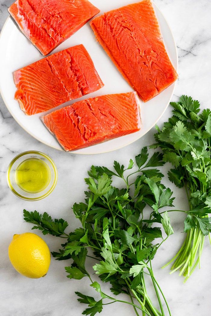 White counter with raw salmon filets on a plate, a bunch of parsley, a bunch of cilantro, a lemon, and a small bowl of olive oil.