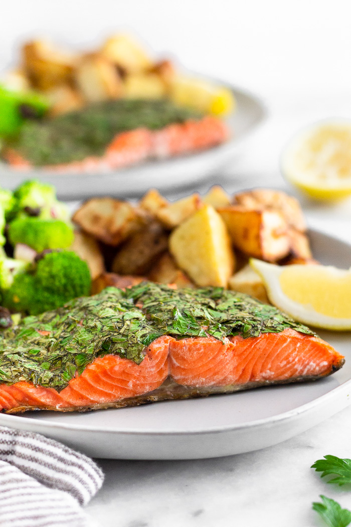 Plate of herb grilled salmon, potatoes, and broccoli salad with a salmon wedge. Behind it is another plate and a lemon wedge.
