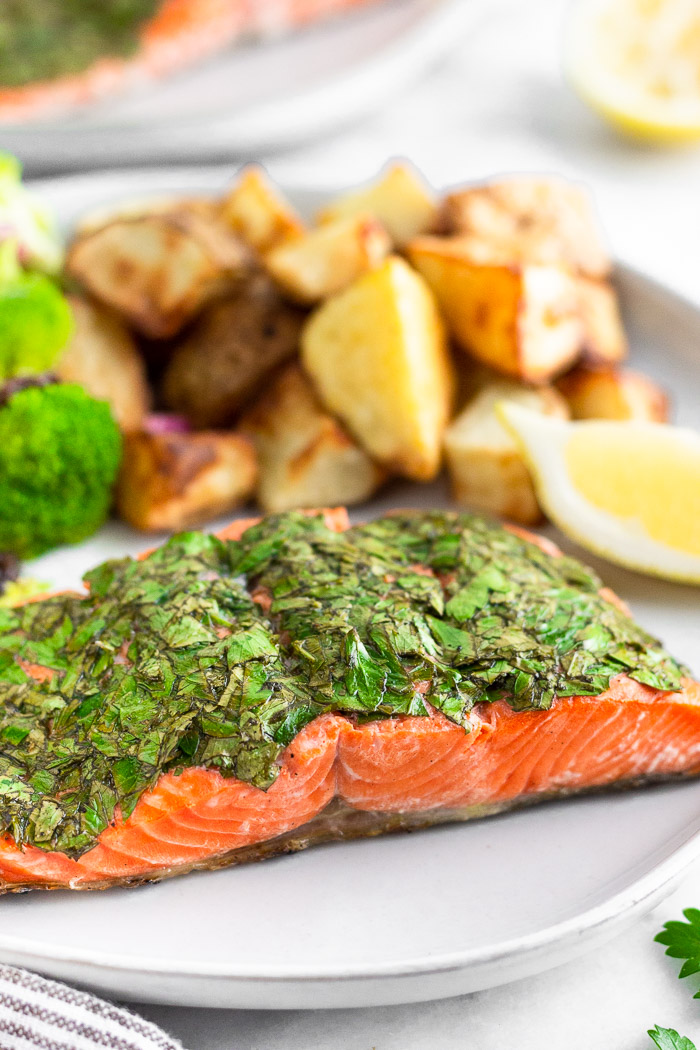 Plate of herb grilled salmon, crispy potatoes, broccoli salad, and a lemon wedge. In the distance is another lemon wedge and another plate of salmon.