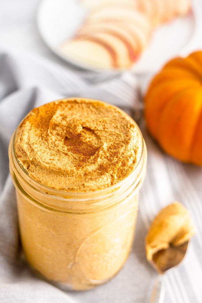 Jar filled with homemade pumpkin pie butter sprinkled with cinnamon. Next to it is a spoon with some on it, a pumpkin, and a plate of sliced apples in the back.