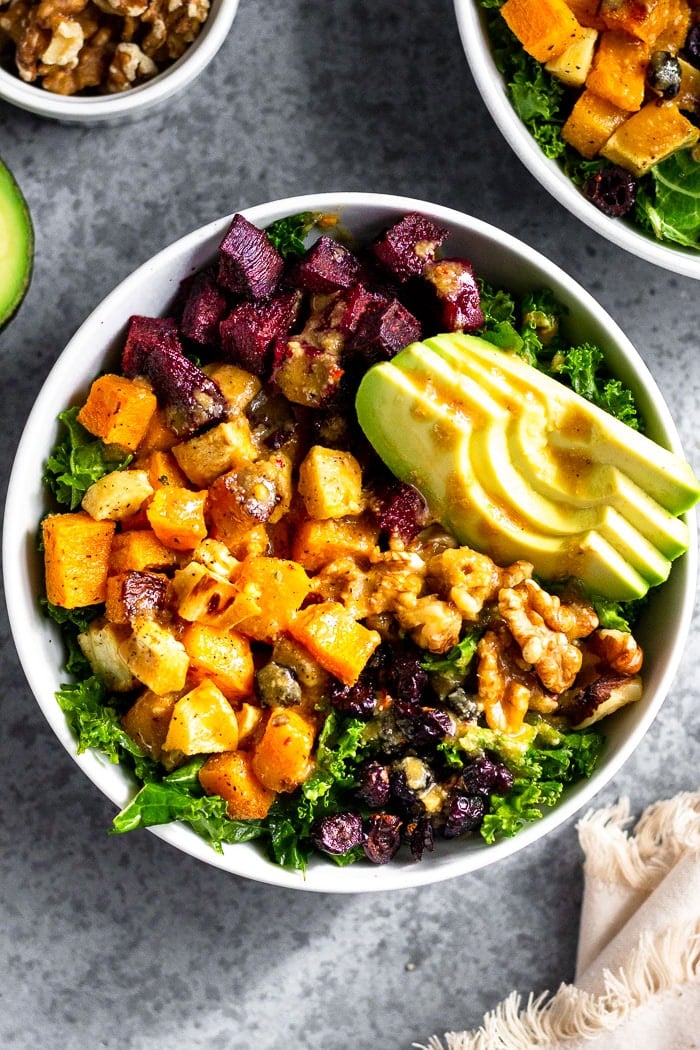 Veggie power bowl with kale, butternut squash, parsnips, beets, avocado, walnuts, and dried cranberries. It is topped with tahini dressing. Next to is is another bowl, half an avocado, bowl of walnuts, and a kitchen towel.