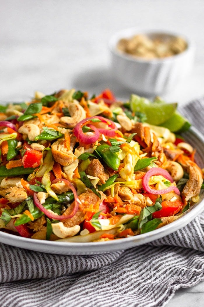 White shallow bowl filled with healthy chicken cabbage salad topped with fresh basil on sitting on a striped towel. A bowl of cashews is behind it.