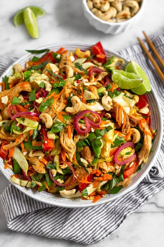 Vietnamese Chicken Cabbage Salad in a white bowl on a blue and white striped towel. Around it are chopsticks, a bowl of cashews, and lime wedges.