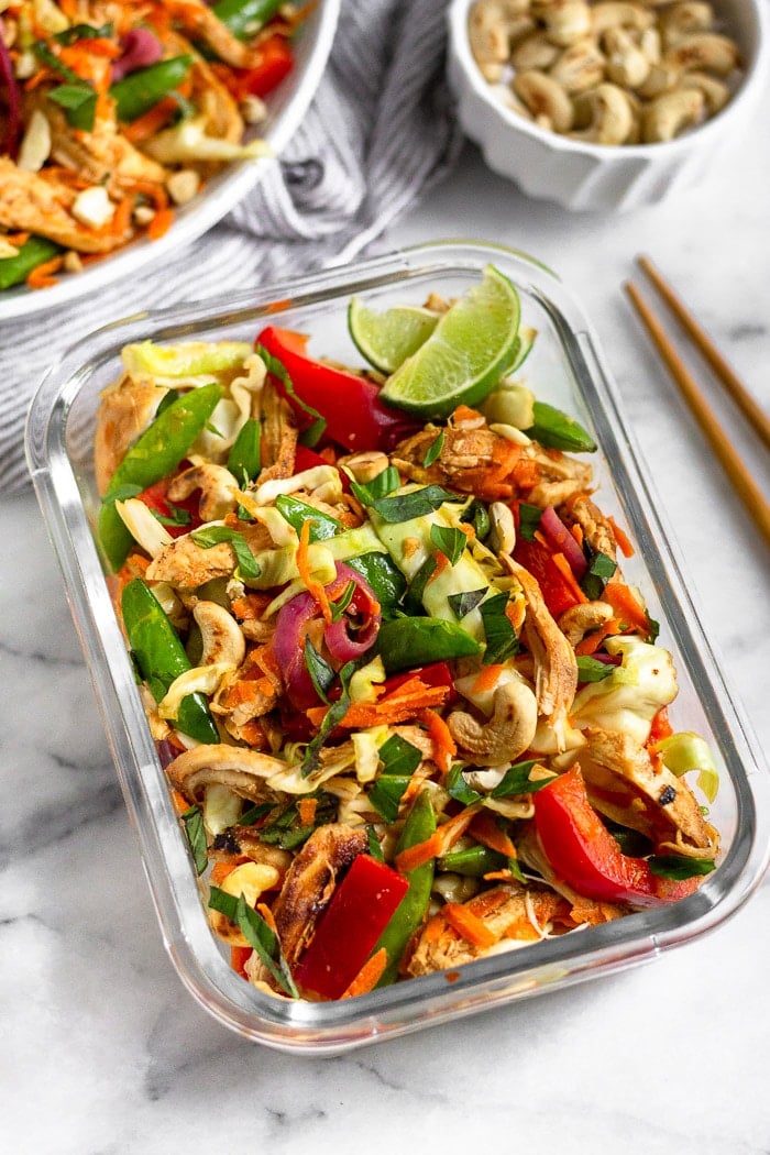 Chicken cabbage salad in a glass meal prep container. Behind it is large bowl of chicken cabbage salad, a bowl of cashews, and chopsticks.