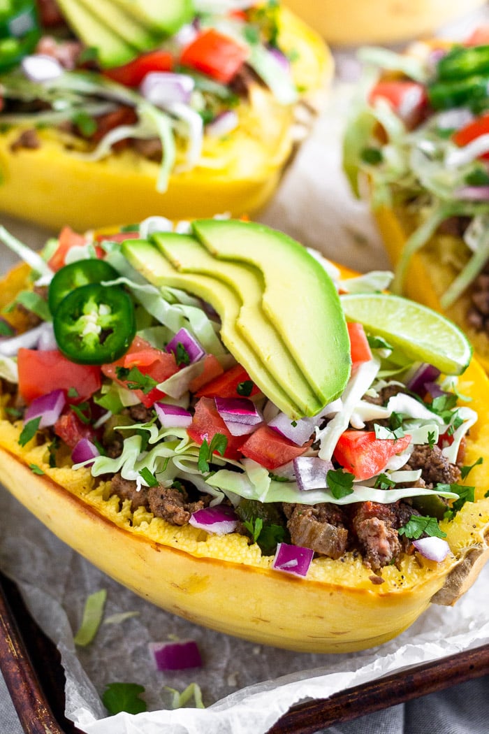 A baking tray with taco stuffed spaghetti squash boats on top that are topped with cabbage, diced tomatoes, avocado, cilantro, and jalapeños.