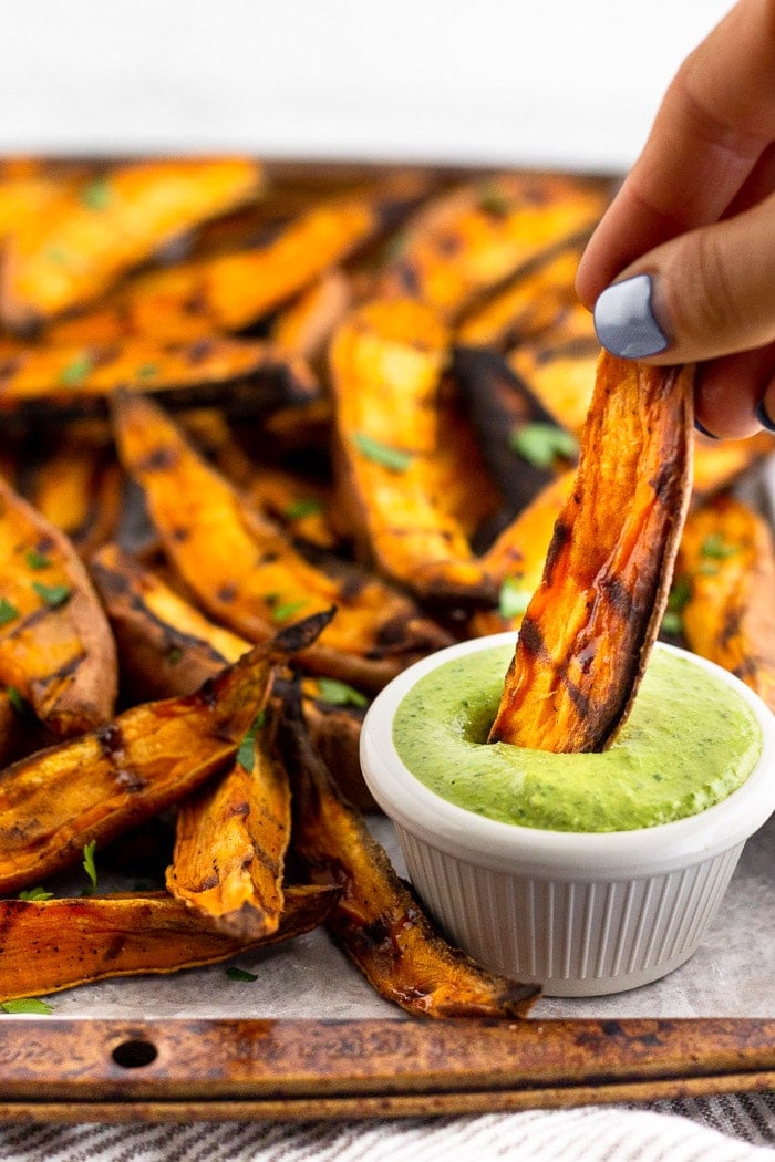 Large sweet of sweet potato wedges with someone holding one and dipping it into a small saucer of green sauce.