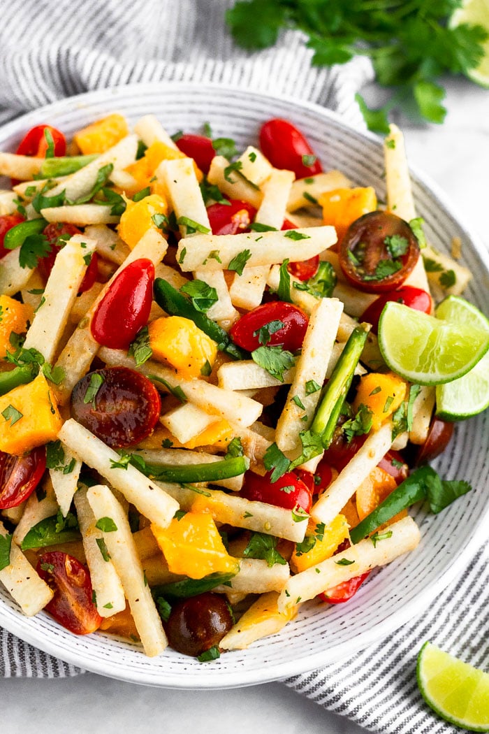 Bowl filled with jicama salad with tomatoes, mango, and jalapeños garnished with lime wedges. It is sitting on a stripped towel with half a lime and cilantro behind it.