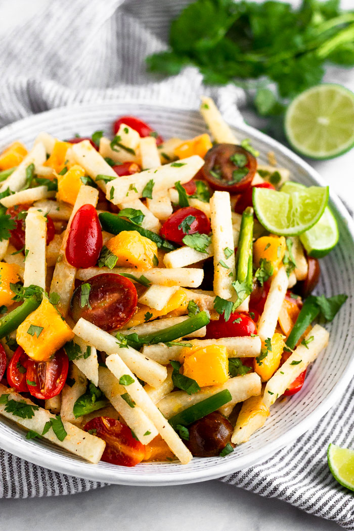 White bowl filled with tomatoes jicama salad with mangos and jalapeños garnished with lime wedges. Behind it is a bunch of cilantro and half a lime.