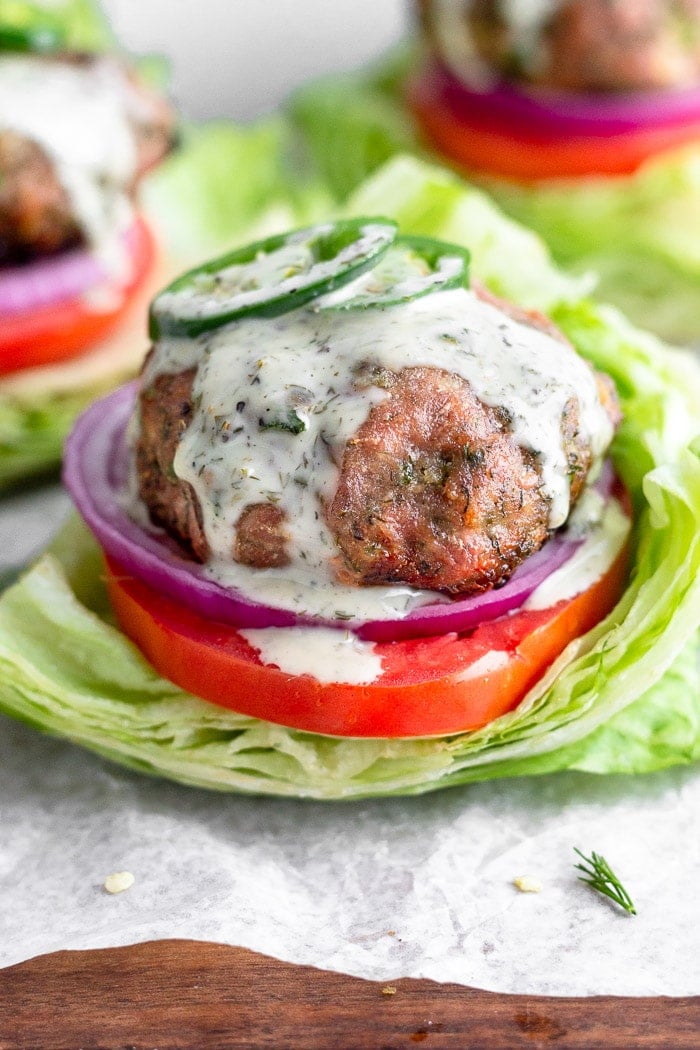 Ranch turkey burger covered in ranch dressing and topped with sliced jalapeños. It is in a lettuce wrap with tomato and red onion. Behind it is more burgers.