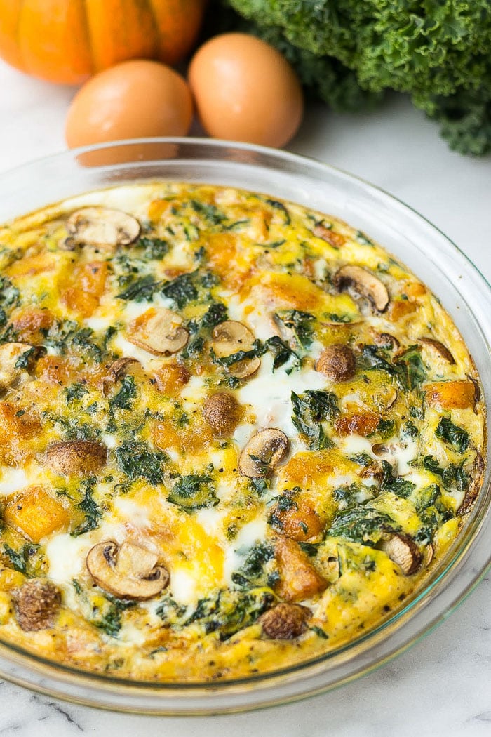 Paleo pumpkin quiche in a pie pan with kale, eggs, and a small pumpkin behind it.