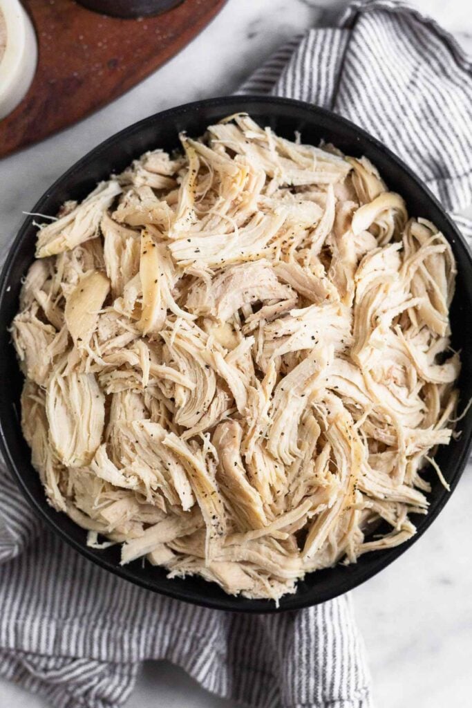 A black bowl filled with shredded chicken sprinkled with pepper. The bowl is on a striped kitchen towel with a wooden dish next to it.
