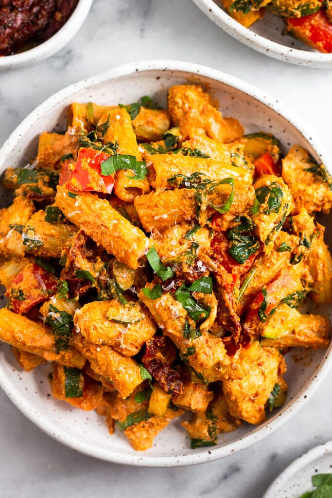 Chicken pasta with sun dried tomatoes in a bowl topped with basil and parmesan cheese. Next to it is another bowl and a bowl of sun-dried tomatoes.