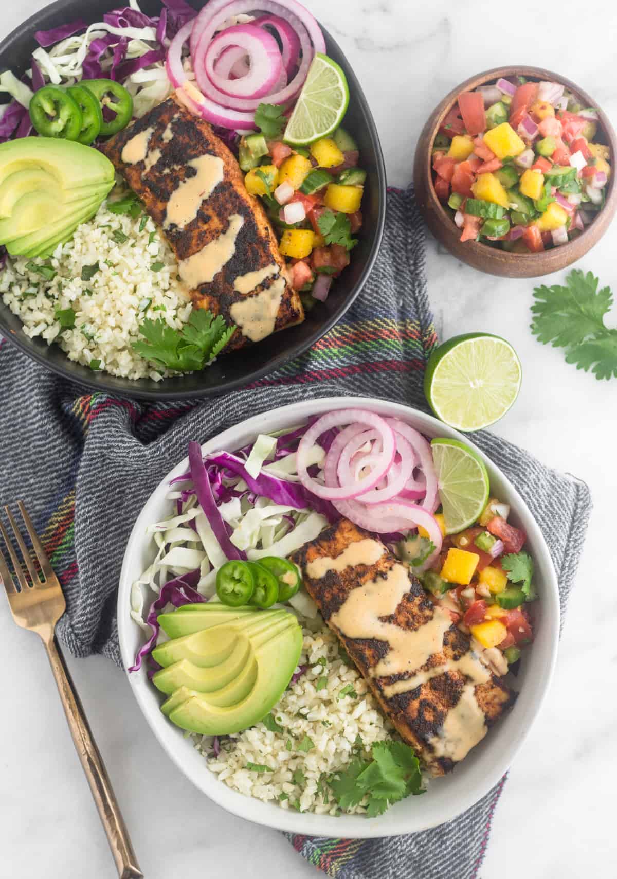 Two Whole30 fish taco bowls on top of a napkin surrounded by a bowl of mango salsa, lime, and cilantro