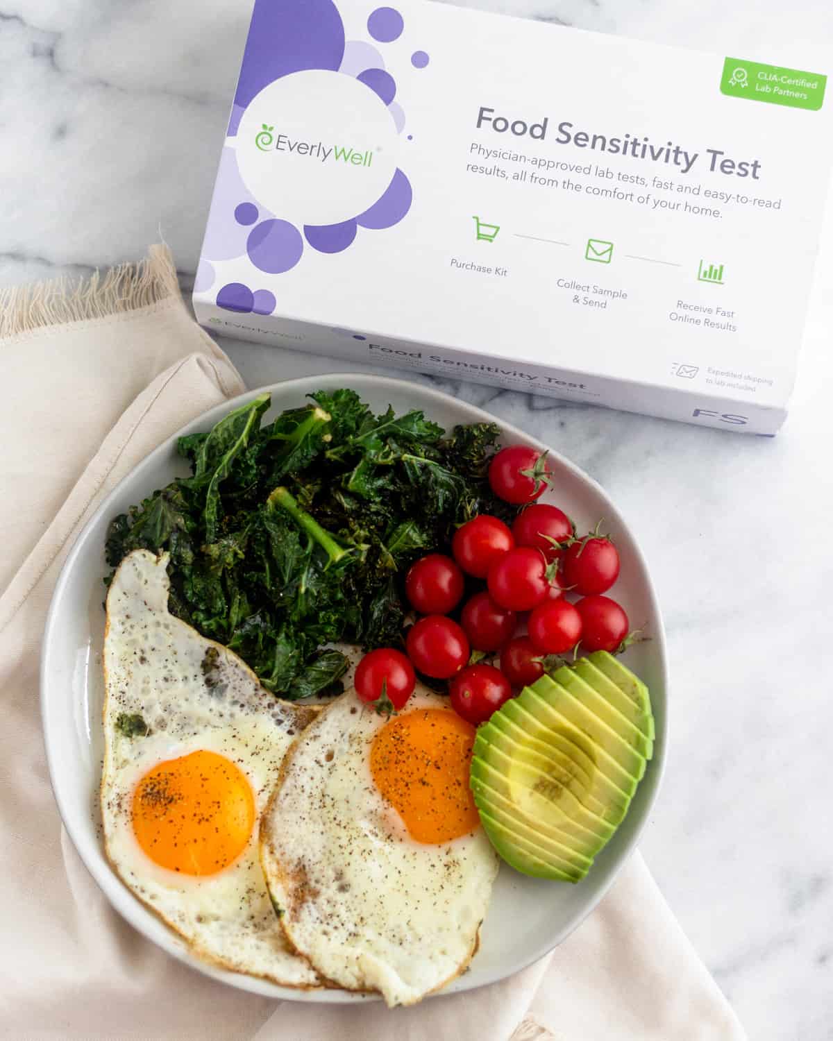 Plate of 2 fried eggs, sautéed kale, cherry tomatoes, and half and avocado on a white counter next to an EverlyWell Food Sensitivity test