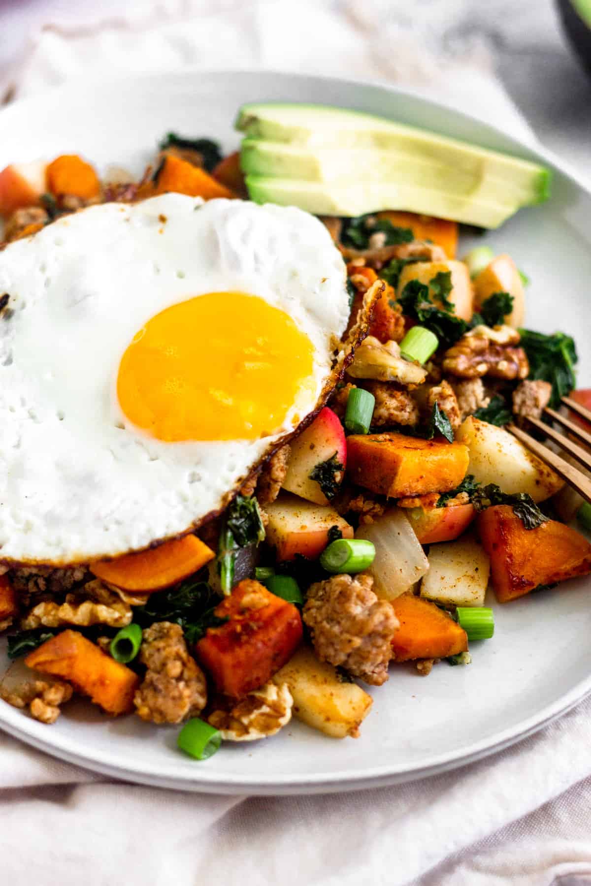 White plate on a tan linen with sweet potato sausage hash topped with a fried egg and sliced avocado.