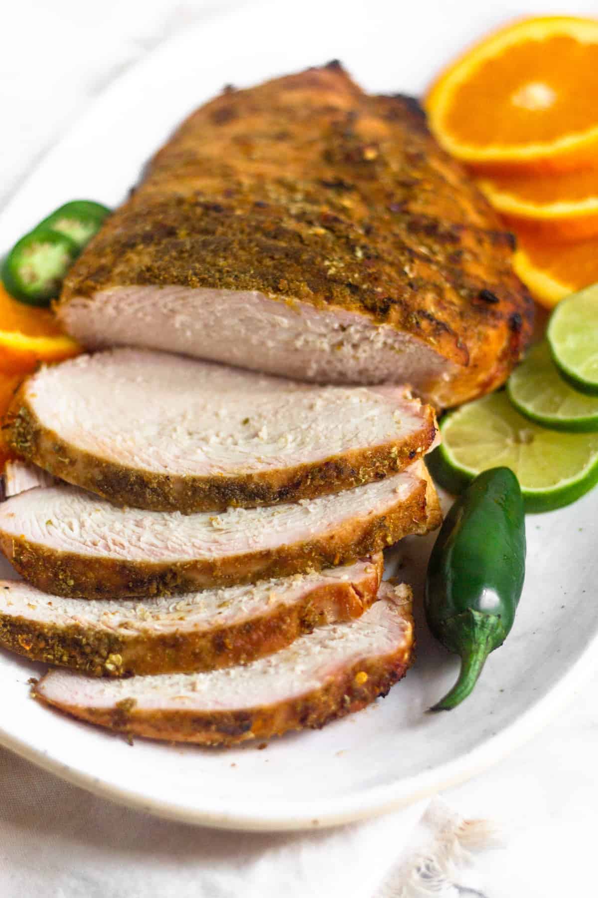 Citrus grilled turkey breast with a few pieces cut laying on a plate next to some sliced limes, oranges, and a jalapeño.