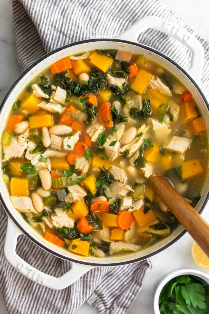 Turkey soup with butternut squash, veggies, kale, and beans in a large white stock pot. There is a wooden spoon in the pot and the pot is sitting on a striped kitchen towel. Next to the pot is half a lemon and a ramekin of fresh herbs.