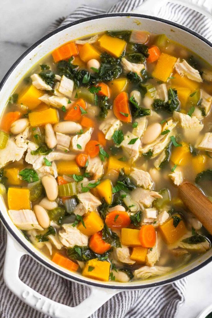 A large pot of leftover turkey soup with butternut squash, kale, veggies, and beans. It is topped with fresh herbs and there is a wooden spoon in the pot.