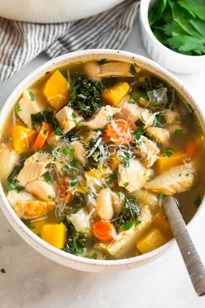A bowl of turkey and butternut squash soup with veggies and beans. There is a spoon in the bowl and it is topped with fresh herbs and grated parmesan cheese. Behind the bowl is a ramekin of fresh herbs and a striped kitchen towel.