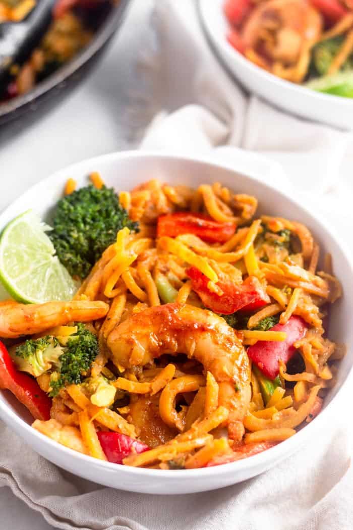 Bowl of paleo and Whole30 Shrimp & Butternut Squash Noodles with Curry Peanut Sauce in a white bowl garnished with a lime wedge. Behind it is another bowl and a large pan filled with more of the noodles. The bowls are sitting on a tan linen.