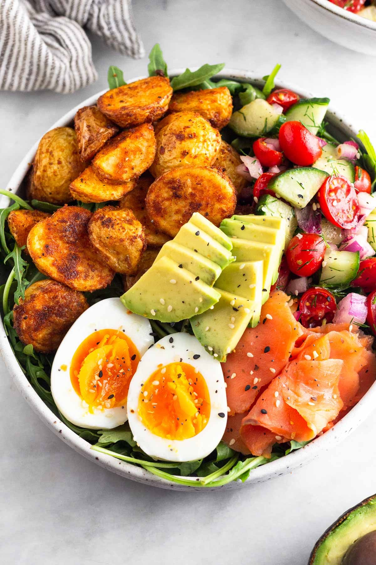 Breakfast bowl filled with greens, roasted potatoes, tomato and cucumber salad, smoked salmon, sliced avocado, and a hard-boiled egg and topped with everything but the bagel seasoning. Next to it is half an avocado.