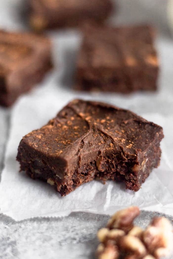 A vegan no bake walnut protein brownie with a bite out of it sitting on a small pieces of white paper. Behind it is more brownies and in front of it is a walnut.