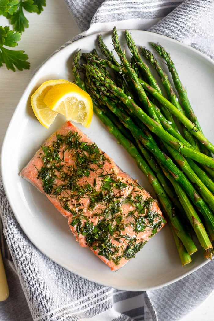 Dill coated salmon and asparagus on a white plate with two lemon wedges next too it. It is on a grey towel with fresh parsley next to it.