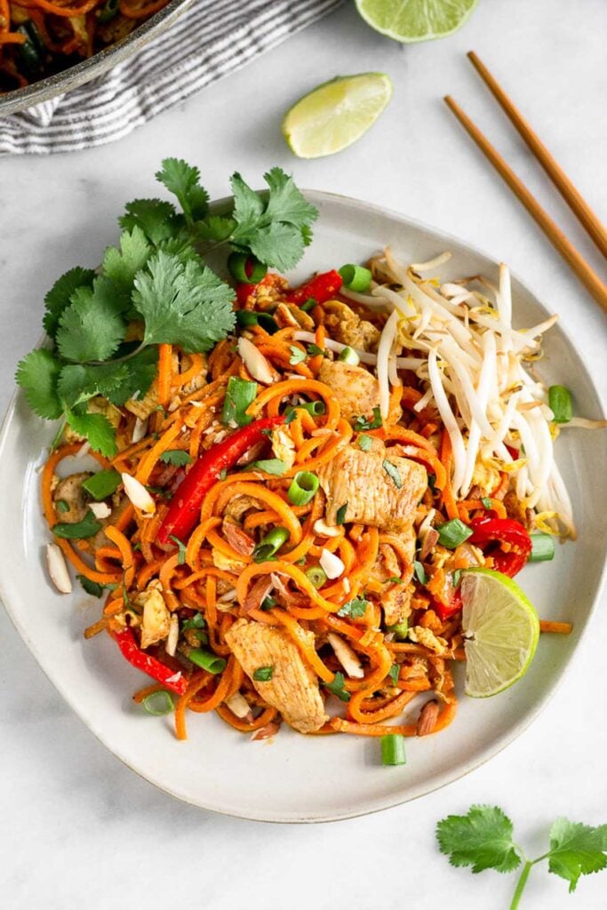 White plate with chicken pad thai made with sweet potato noodles topped with bean sprouts, cilantro, and a lime wedge. Next to it is chopsticks, lime wedges, and cilantro.