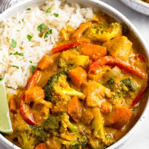 Large white bowl filled with Thai coconut chicken curry and white rice. It is garnished with a lime wedge and fresh cilantro. Around it is another bowl of curry.