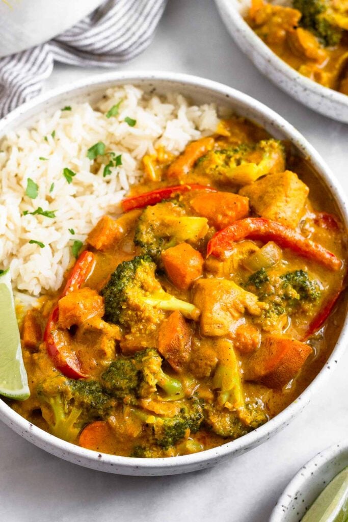 Large white bowl filled with Thai coconut chicken curry and white rice. It is garnished with a lime wedge and fresh cilantro. Around it is another bowl of curry.