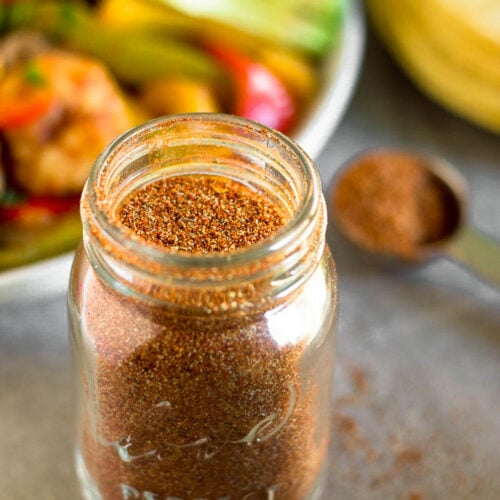A jar filled with homemade taco seasoning. Behind it is a tablespoon full of seasoning, a plate of fajitas, and some tortillas.