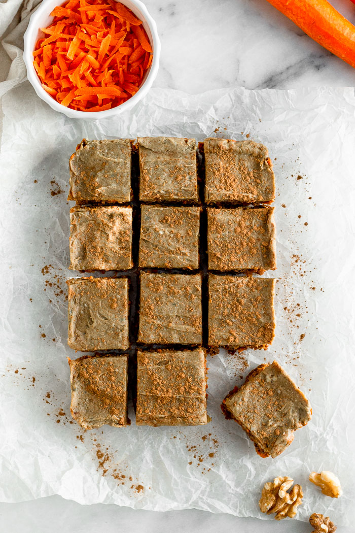 Overhead shot of vegan carrot cake protein bars that are cut into 12 squares. Ones of the square is pulled away from the others. Surrounded them are walnuts and carrots.