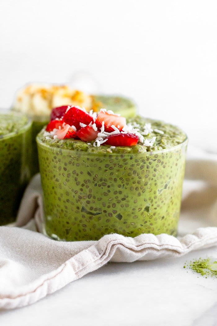 A glass cup filled with matcha chia pudding topped with fresh chopped strawberries and coconut shreds. Behind and next to it is more jars of chia pudding.