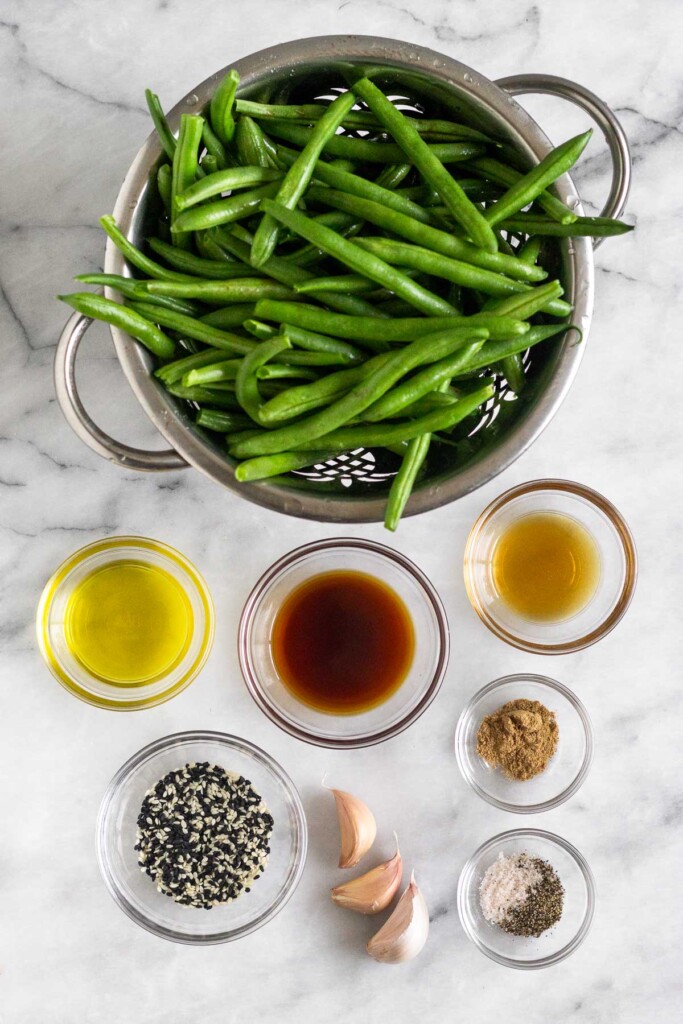 White marble counter with a colander of green beans, a bowl of sesame oil, a bowl of ground ginger, a bowl of salt and pepper, 3 cloves of garlic, a bowl of sesame seeds, a bowl of coconut aminos, and a bowl of olive oil.