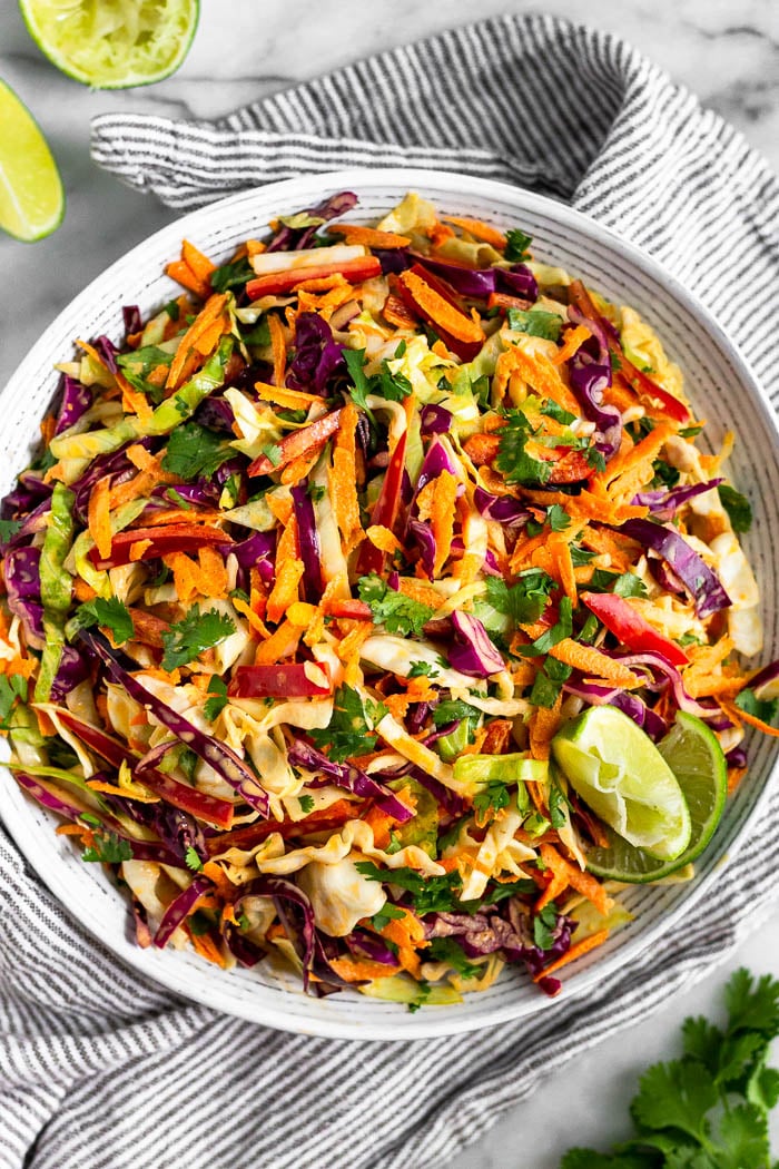 Large bowl of chipotle Mexican coleslaw with lime wedges on the side of the bowl. It is stilling on a stripped towel with lime wedges and cilantro around it.