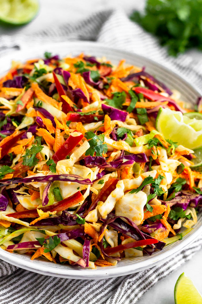 A bowl of spicy paleo Mexican coleslaw garnished with lime wedges sitting on a stripped towel. Around it is a bunch of cilantro and lime wedges.