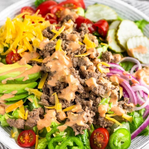 Burger salad filled with lettuce, ground beef, tomatoes, pickles, onions, avocado, cheese, and topped with special sauce. Behind it is a jar of special sauce and a plate of more toppings.