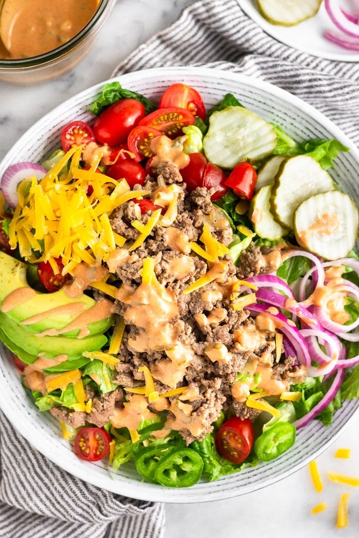 Overhead view of a cheeseburger salad with lettuce, tomato, ground beef, onions, pickles, jalapeños, avocado, and cheese all topped with special sauce. It is surrounded by a jar of special sauce, a plate of more toppings, and few shreds of cheese next to the bowl.
