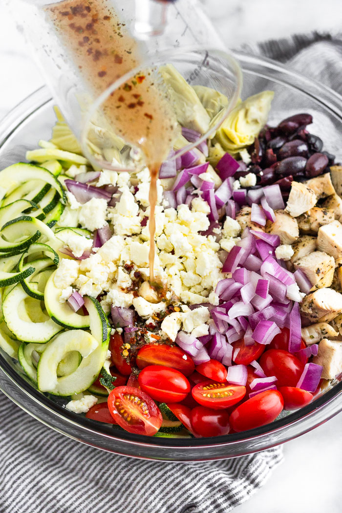 Dressing being poured overtop a large bowl filled with zucchini noodles, cherry tomatoes, red onion, chopped chicken, olives, and artichokes hearts.