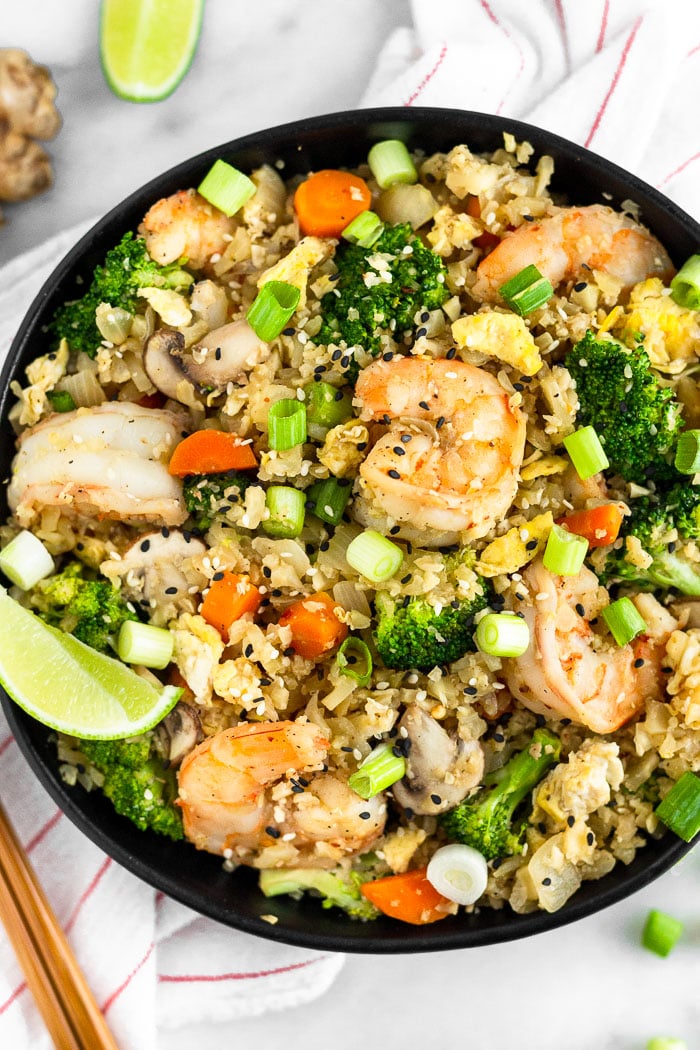 Large bowl of paleo cauliflower shrimp fried rice garnished with green onions and a lime wedge. The bowl is sitting on a stripped towel with chopsticks, ginger, a lime wedge, and green onions around it.