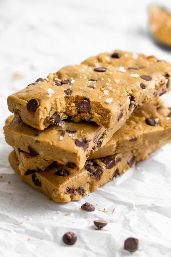 A stack of three peanut butter chocolate chip protein bars on top of each other. The one on top has a bite taken out of it. In front of the stack are a few mini chocolate chips.