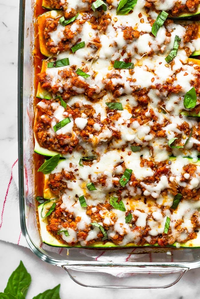 Healthy zucchini pizza boats in a large glass dish toped with fresh basil and red pepper flakes. The pan is sitting on a stripped towel with basil leaves next to it.