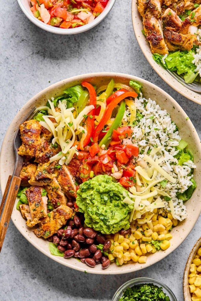 Overhead shot of a copycat chipotle chicken burrito bowl with cilantro lime rice, corn, black beans, sliced seasoned chicken, lettuce, sautéed peppers and onions, shredded cheese, pico de gallo, and guacamole. There is a fork in the bowl as well. Around the bowl is a bowl of corn, a bowl of cilantro, a bowl of pico de gallo, and another burrito bowl.
