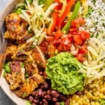 Overhead shot of a chicken burrito bowl with cilantro lime rice, corn, black beans, guacamole, sliced seasoned chicken, lettuce, shredded cheese, fajita veggies, and pico de gallo. A fork is in the bowl as well.