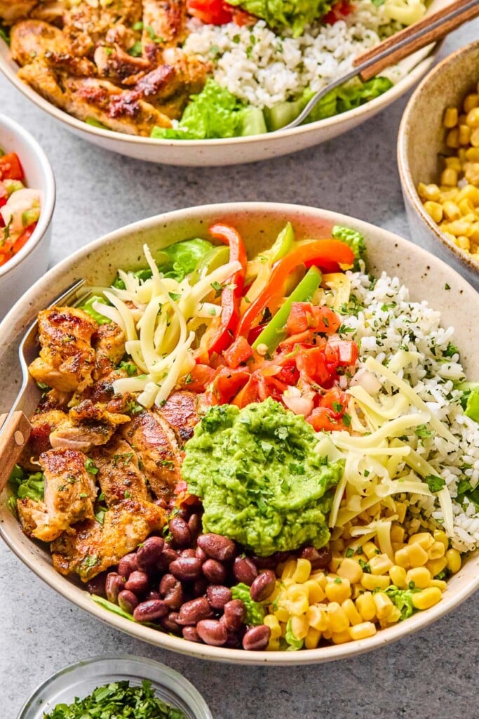 Chicken burrito bowls with seasoned chicken, fajita veggies, cilantro lime rice, black beans, corn, cheese, salsa, and guacamole. Around the bowl is a small bowl of cilantro, a bowl of salsa, another burrito bowl, and a bowl of corn.