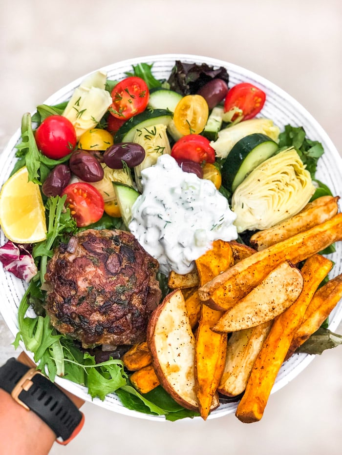Burger salad with tomatoes, cucumbers, artichokes, olives, French fries, a lamb burger, and homemade tzatziki showing how you can enjoy food freedom with nutrition coaching.