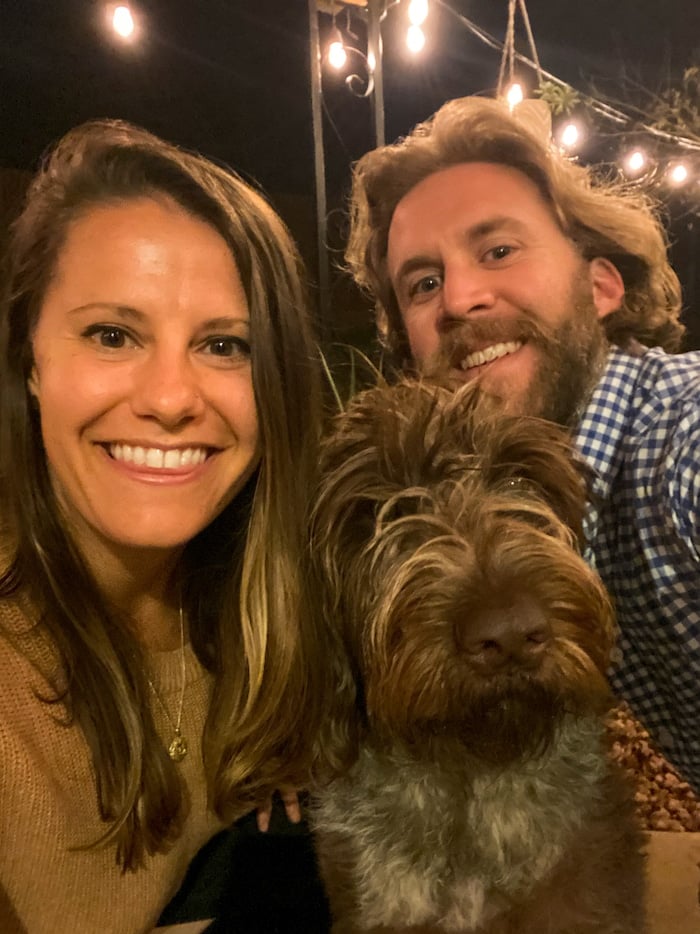 Woman, man, and a dog taking a selfie at night with lights on in the background.