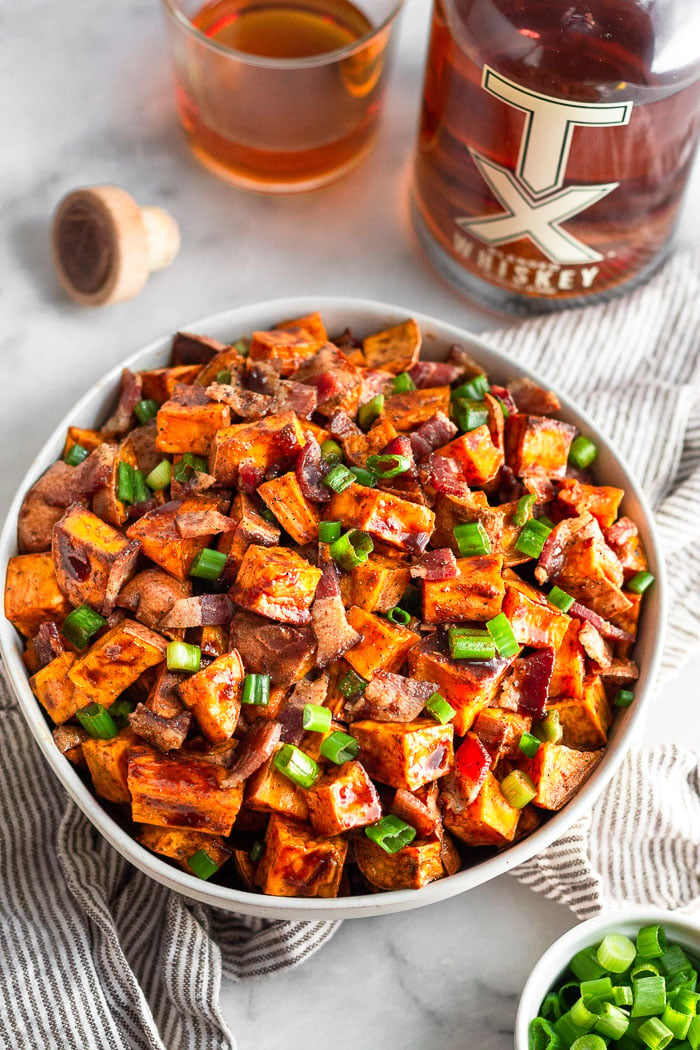 Bowl of whiskey glazed sweet potatoes topped with bacon and green onion. The bowl is sitting on a striped towel with a bowl of green onions, bottle of whiskey, a cup of whiskey, and the bottle's cork next to it.