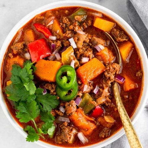 Bison chili in a bowl topped with cilantro and jalapeños with a spoon in it. Another it are some lime wedges, another bowl of chili, and dish of cilantro.